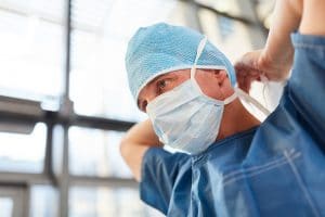 male critical care nurse in blue scrubs tying his face mask onto his face