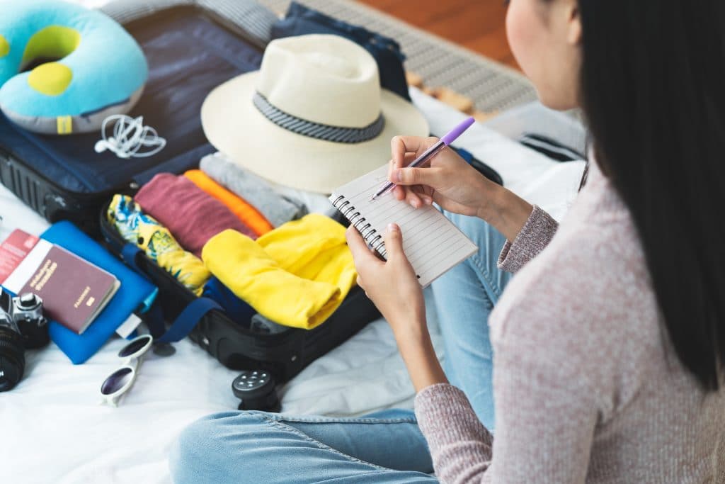 Preparing suitcase for summer vacation trip. Young woman checking accessories and stuff in luggage on the bed at home before travel.