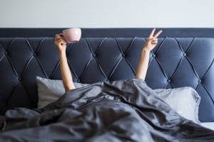 woman just waking up in bed with arms up, one hand with coffee mug and the other a peace sign