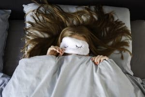 young woman sleeping in her bed with an eye mask and covers over her mouth.