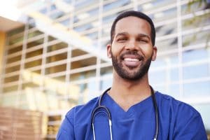 black male nurse standing outside hospital in blue scrubs with a smile on his face